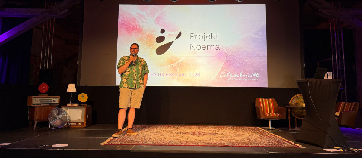 Eine Vortragsb&uuml;hne gem&uuml;tlich eingerichtet wie ein Wohnzimmer der 1960er Jahre. Lutz Schmitt steht auf der B&uuml;hne in Hawaii-Hemd und Shorts und pr&auml;sentiert Projekt Noema.